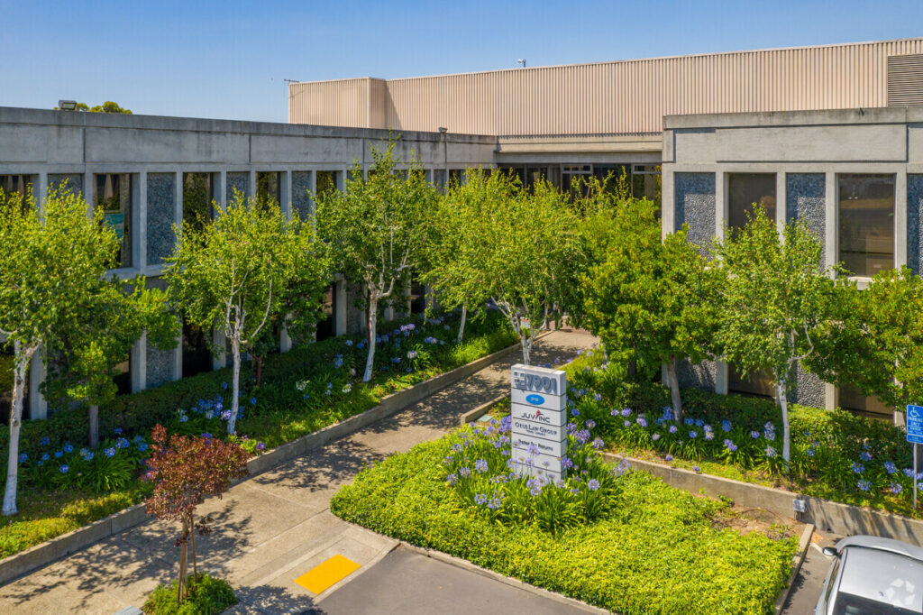 Aerial of entryway landscaping of 7901 Oakport. Delicate green trees and blooming purple flowers line windowed walls.