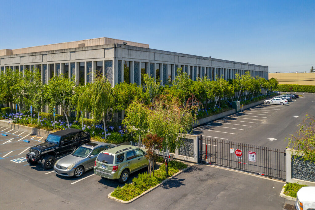 Aerial of 7901 Oakport. Delicate green trees and blooming purple flowers line windowed walls. Surrounded by gated parking lot.