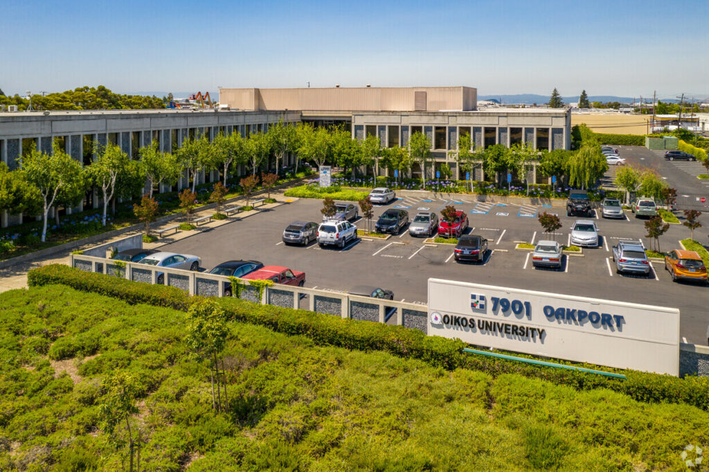 Aerial of 7901 Oakport. Delicate green trees and blooming purple flowers line windowed walls. Surrounded by gated parking lot.