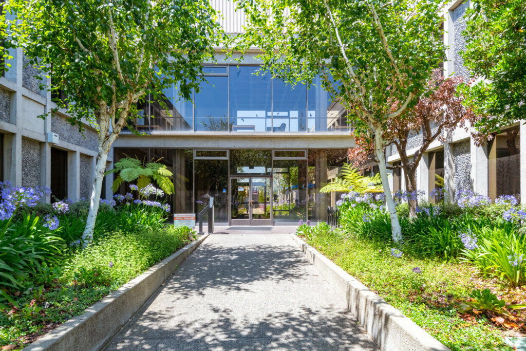 Entryway landscaping of 7901 Oakport. Delicate green trees and blooming purple flowers line windowed walls.