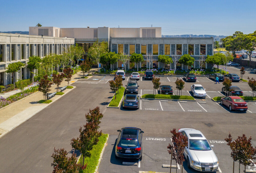 Aerial of 7901 Oakport. Delicate green trees and blooming purple flowers line windowed walls. Surrounded by gated parking lot.